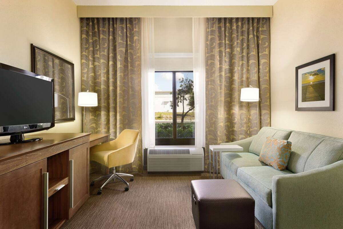 A hotel room with a flat-screen TV, a desk, a chair, a sofa, and a window with curtains.