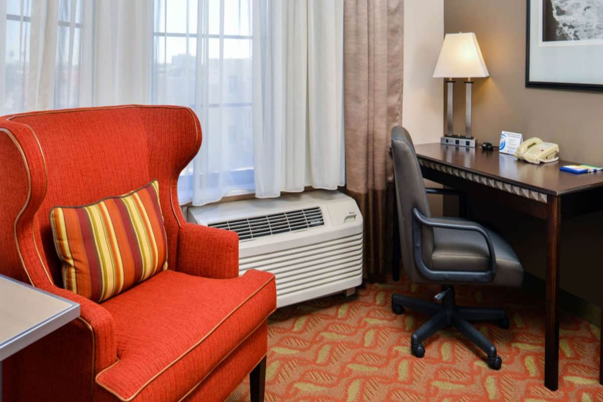 Une chambre d’hôtel avec un fauteuil rouge, une chaise de bureau noire, un bureau avec une lampe, un téléphone et un tableau encadré au mur.