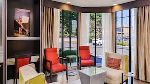 Een hotellobby met een zithoek, bestaande uit rode en beige stoelen, een glazen salontafel en een kamerbreed tapijt. Grote ramen bieden uitzicht op de straat.