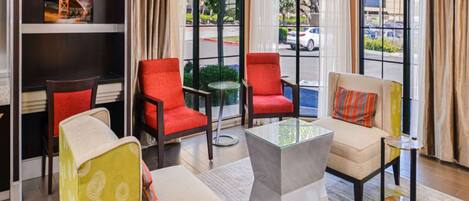 A hotel lobby with a seating area featuring red and beige chairs, a glass coffee table, and a patterned rug. Large windows provide views of the street outside.