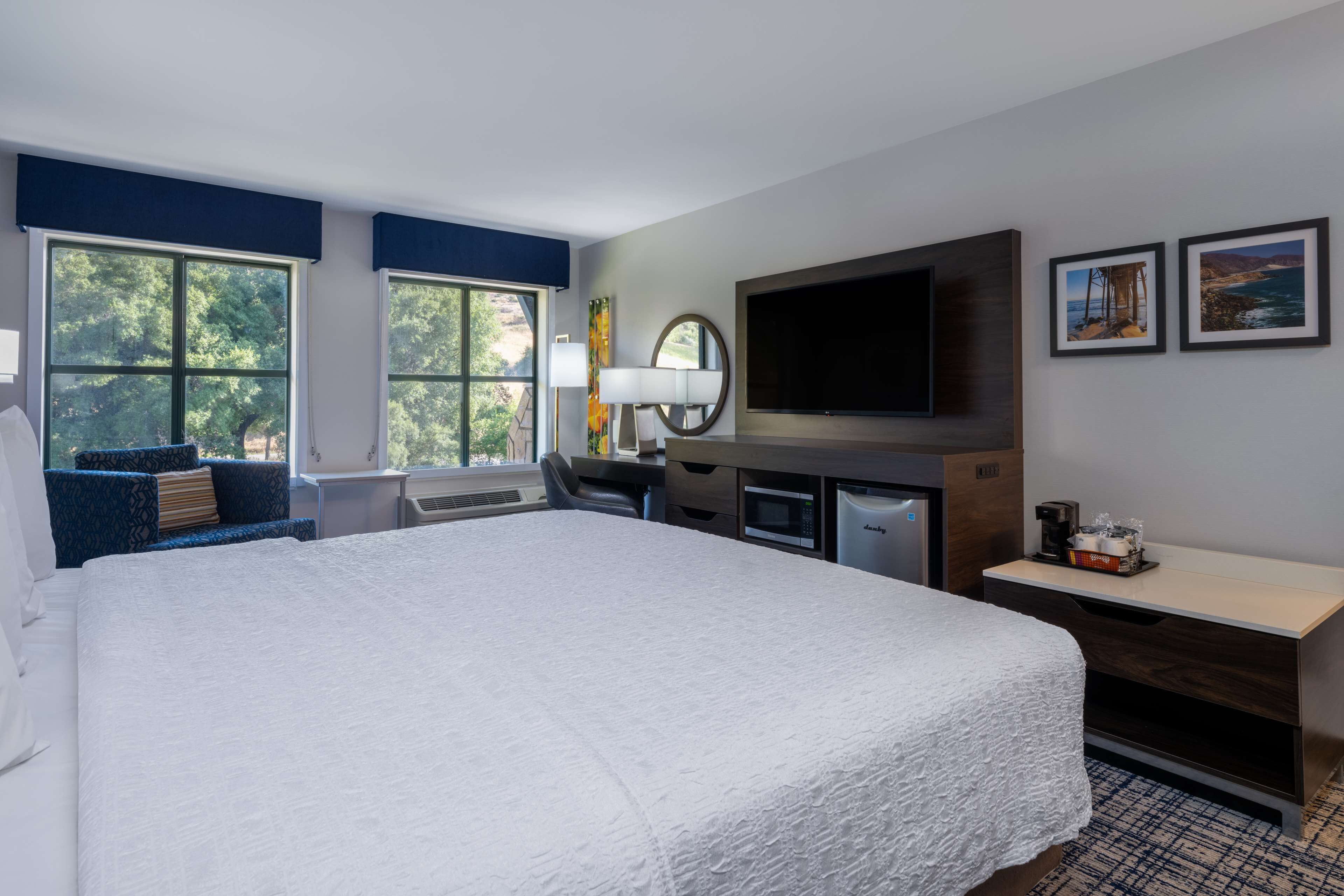 Une chambre d’hôtel avec un grand lit, un bureau doté d’un téléviseur à écran plat, un four à micro-ondes et une machine à café. La chambre dispose de deux fenêtres avec des rideaux bleus, d’un téléviseur fixé au mur et de tableaux encadrés accrochés au mur.