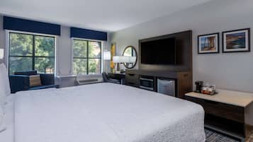 Une chambre d’hôtel avec un grand lit, un bureau doté d’un téléviseur à écran plat, un four à micro-ondes et une machine à café. La chambre dispose de deux fenêtres avec des rideaux bleus, d’un téléviseur fixé au mur et de tableaux encadrés accrochés au mur.