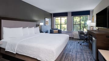 A hotel room with a large bed, a desk, a chair, a TV, and a window with a view of trees.