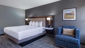 Quarto de hotel com uma cama grande, uma mesa de cabeceira com abajur, uma cadeira e um quadro na parede.