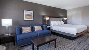 Un cuarto de hotel con una cama grande, un sofá azul, una mesita, una mesita de noche con una lámpara y un cuadro enmarcado en la pared.