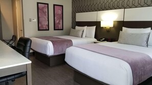 Ein Hotelzimmer mit zwei Betten, einem Schreibtisch und einem Stuhl. An der Wand hängen zwei gerahmte Bilder, ein Nachttisch mit einer Lampe und ein Telefon.
