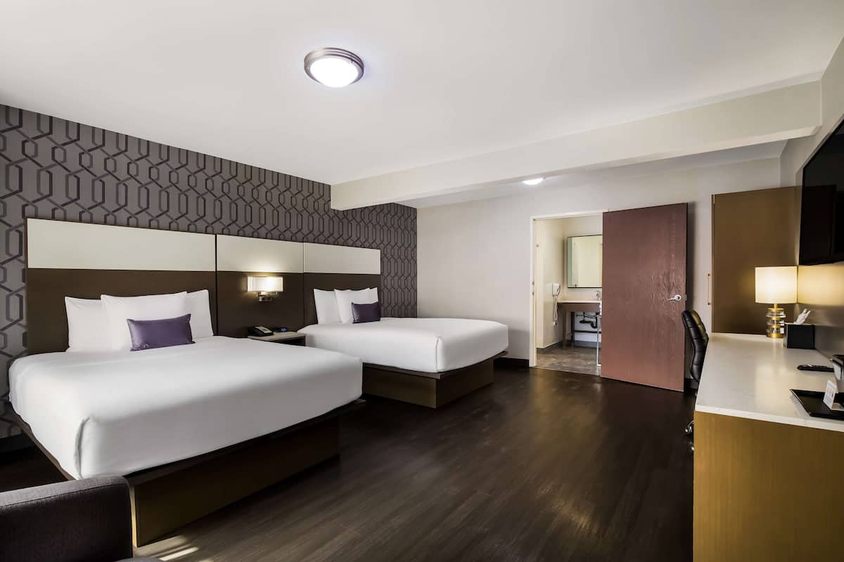 A hotel room with two beds, a desk, a mirror, and a bathroom visible through an open door.