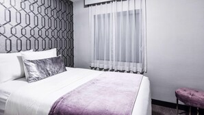 Habitació d'hotel ben ordenada amb un llit, una finestra amb cortines lleugeres, un mur amb un disseny i una otomana morada petita.