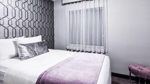 Ein ordentlich eingerichtetes Hotelzimmer mit einem Bett, einem Fenster mit durchscheinenden Vorhängen, einer Wand mit Muster und einem kleinen violetten Hocker.