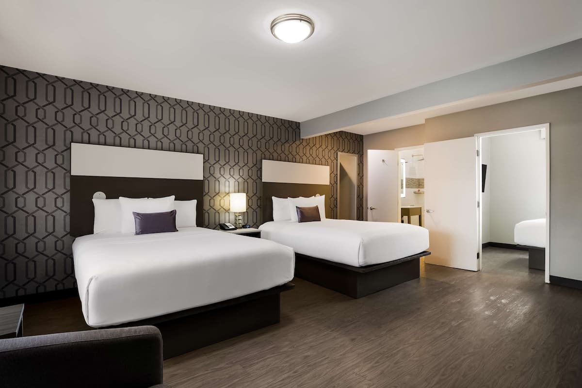 A hotel room with two beds, a nightstand with a lamp, a wooden floor, and a patterned wall.
