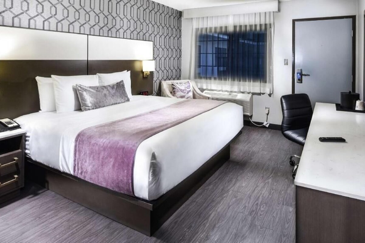 A hotel room with a bed, bedside table, chair, desk, and a TV.