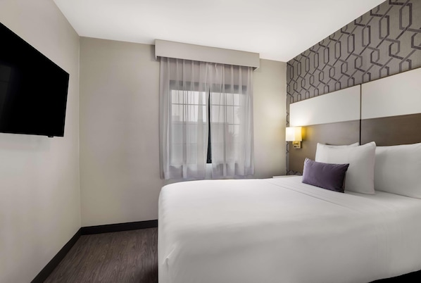 Ein Hotelzimmer mit einem großen Bett, einem flachbildfernseher an der Wand, einem Fenster mit durchsichtigen Vorhängen und einem gemusterten Kopfteil.