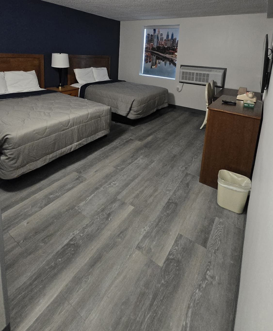 Hotelkamer met twee bedden, een houten hoofdbord, een nachtkastje met een lamp, een raam met uitzicht op de stad, een heater en een klein bureau met een afvalbak.
