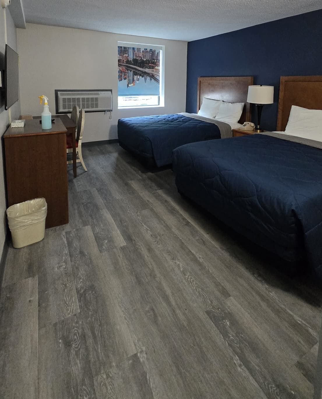 A hotel room with two beds, a desk, a chair, a trash bin, and a window with a cityscape view.