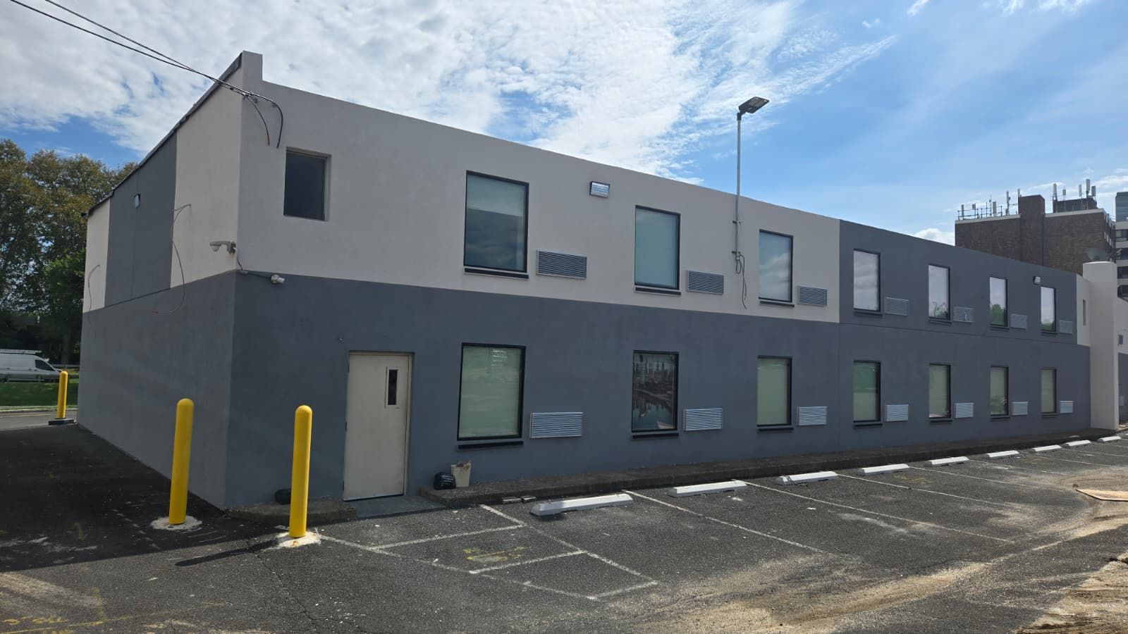 A modern building with a flat facade, multiple windows, and a parking lot with yellow bollards.