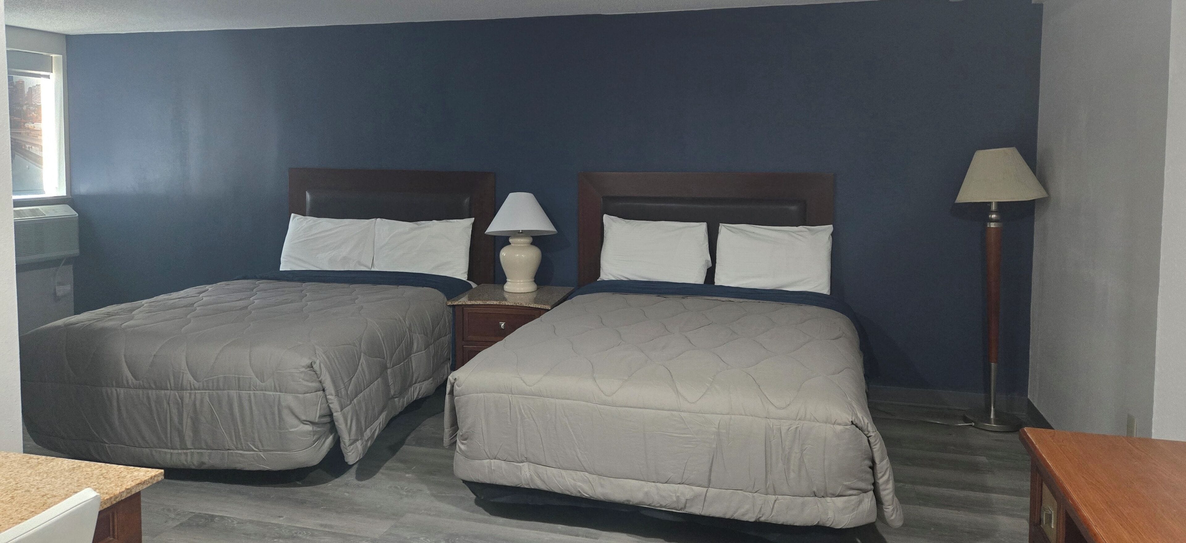 A hotel room with two beds, a nightstand, a lamp, and a window with a city view.