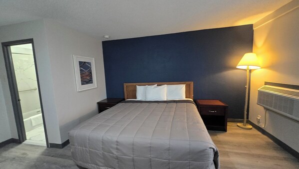 A hotel room with a bed, a nightstand, a bedside lamp, a framed picture on the wall, and a door leading to a bathroom. - Roosevelt Inn (Philadelphia)