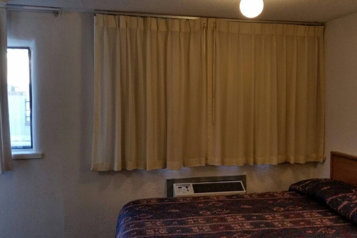 A hotel room with a bed, a window with curtains, and a wall-mounted air conditioning unit.