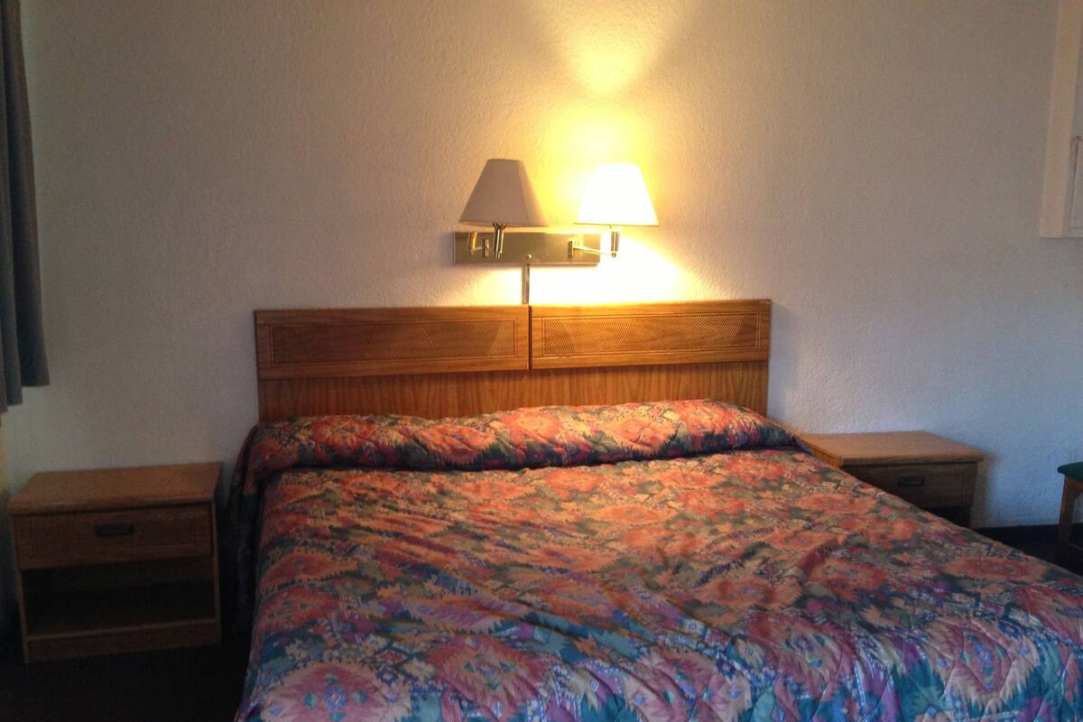 A hotel room with a wooden headboard, a patterned bedspread, two bedside tables, and a wall-mounted lamp.