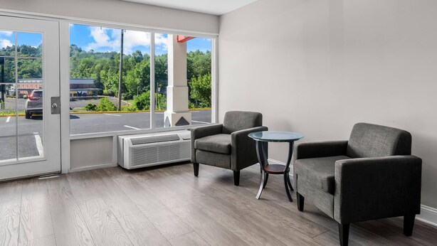 Habitación de hotel moderna con un amplio ventanal corredero que da a un estacionamiento, dos sillones grises, una pequeña mesa redonda y un equipo de aire acondicionado empotrado.