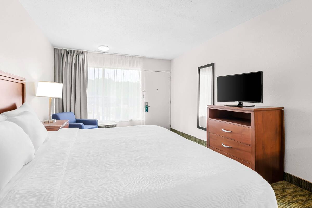 A hotel room with a large bed, a television on a wooden cabinet, a desk with a lamp, a chair, and a window with curtains.