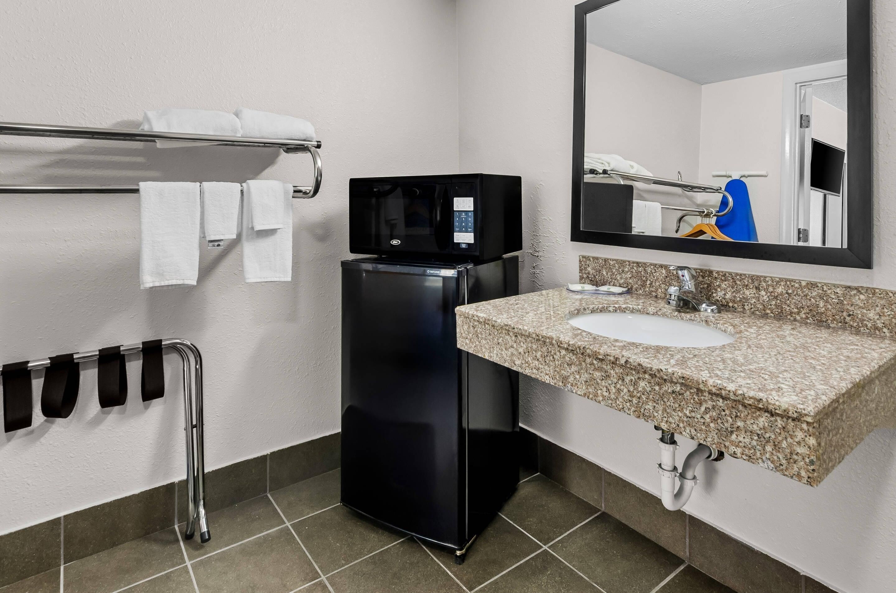 A compact hotel bathroom with a sink, mirror, towel rack, and a black refrigerator.