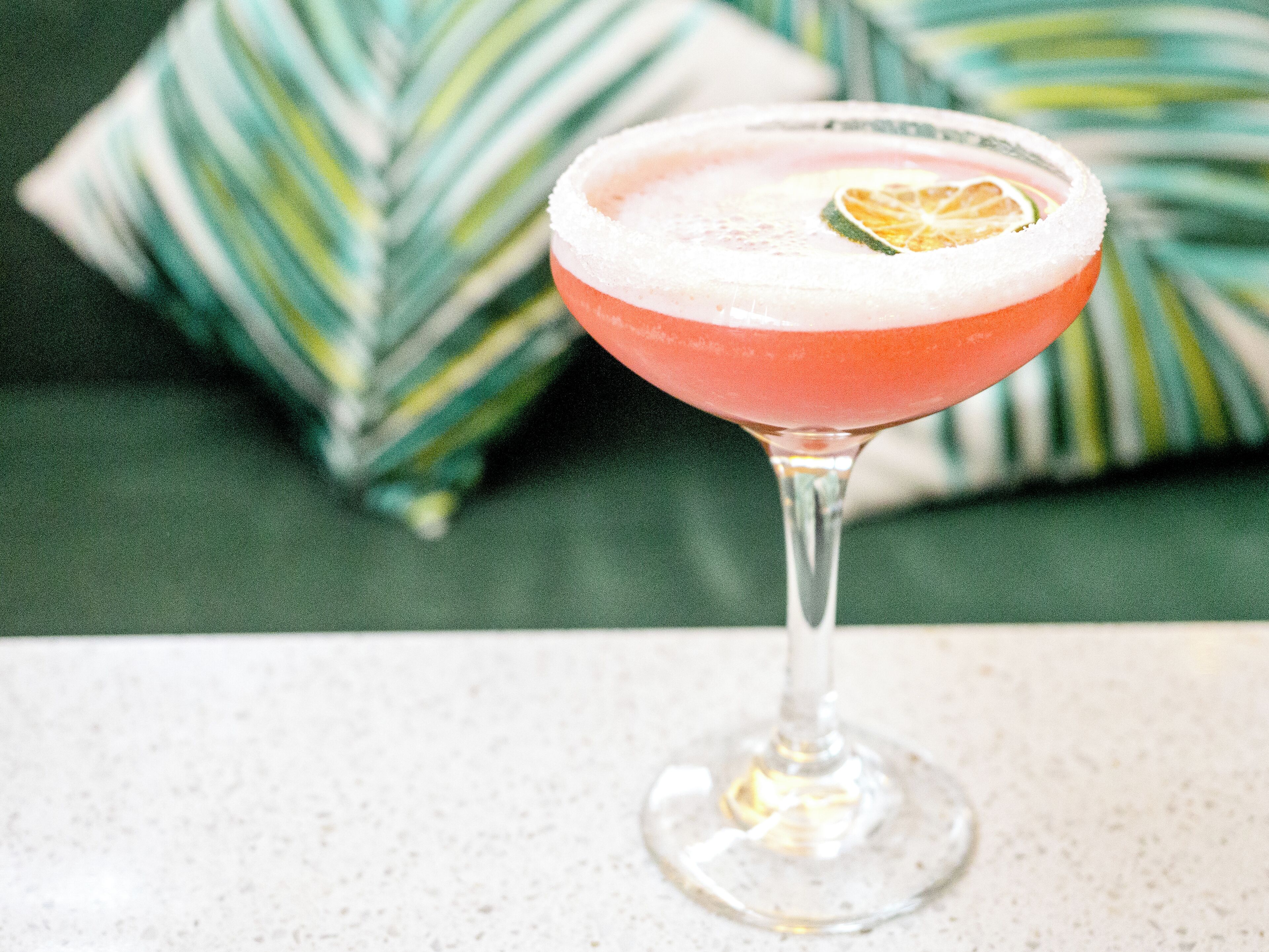 A cocktail in a stemmed glass with a salted rim, garnished with a slice of lime, on a speckled countertop.