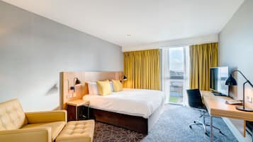A hotel room with a large bed, a desk with a computer, a chair, and a view of the city through the window.