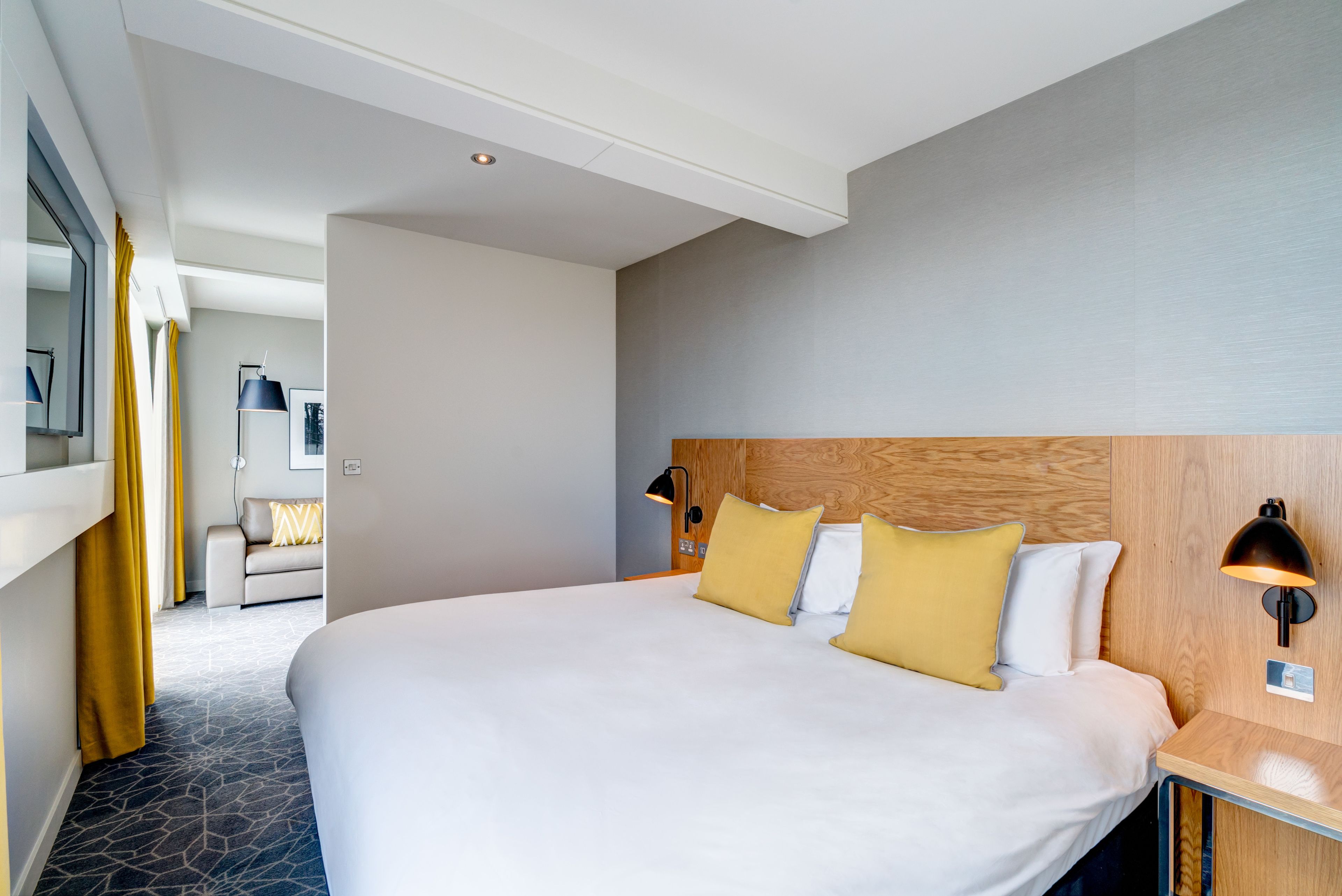 A hotel room with a large bed, a wooden headboard, a bedside table with a lamp, and a view of a hallway with a sofa and curtains.