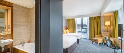 A hotel room with a large bed, a desk, a chair, a bathroom with a sink and bathtub, and a view of the city.