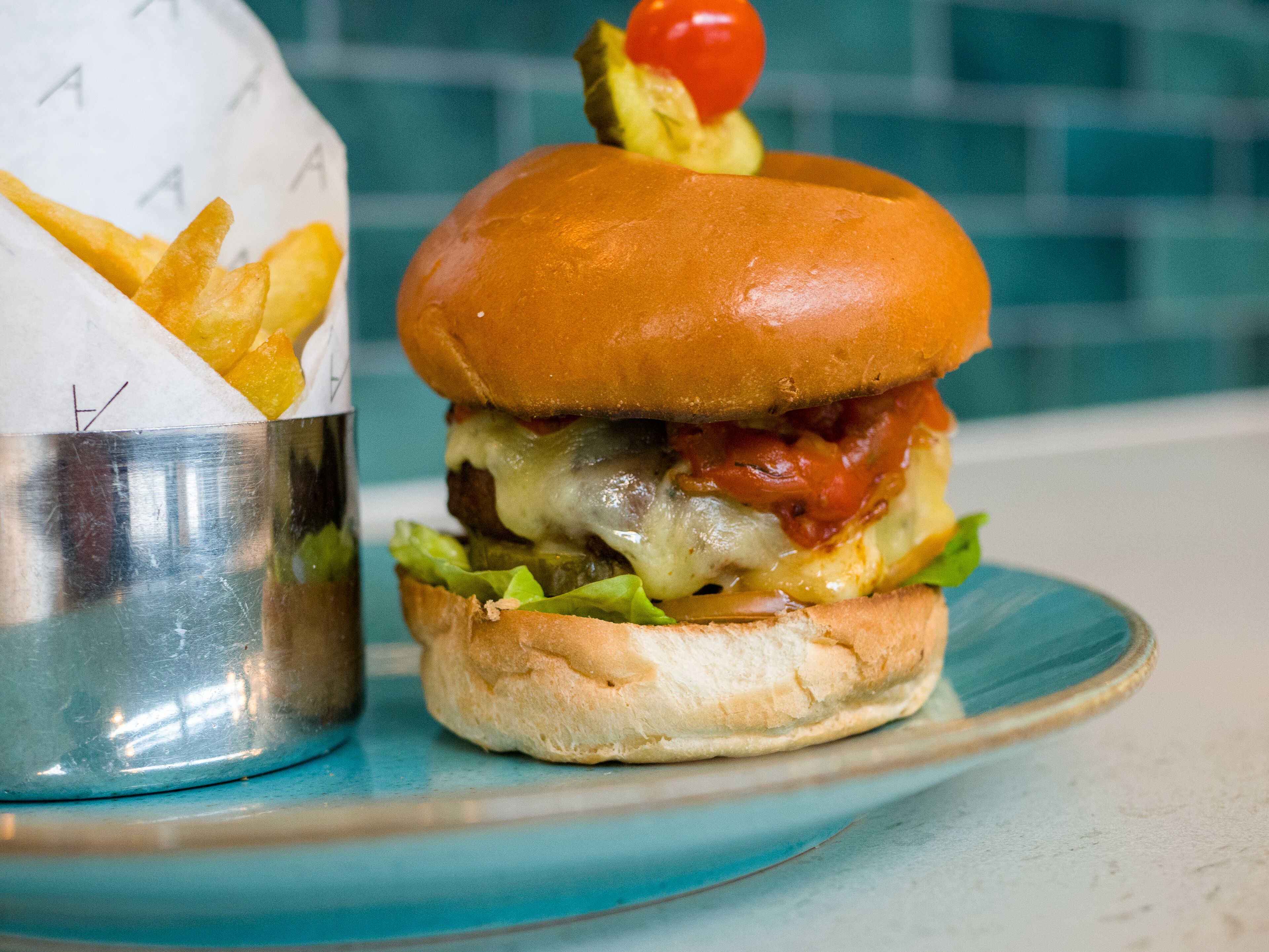 Una hamburguesa con tomate encima, una porción de papas fritas y un recipiente metálico.