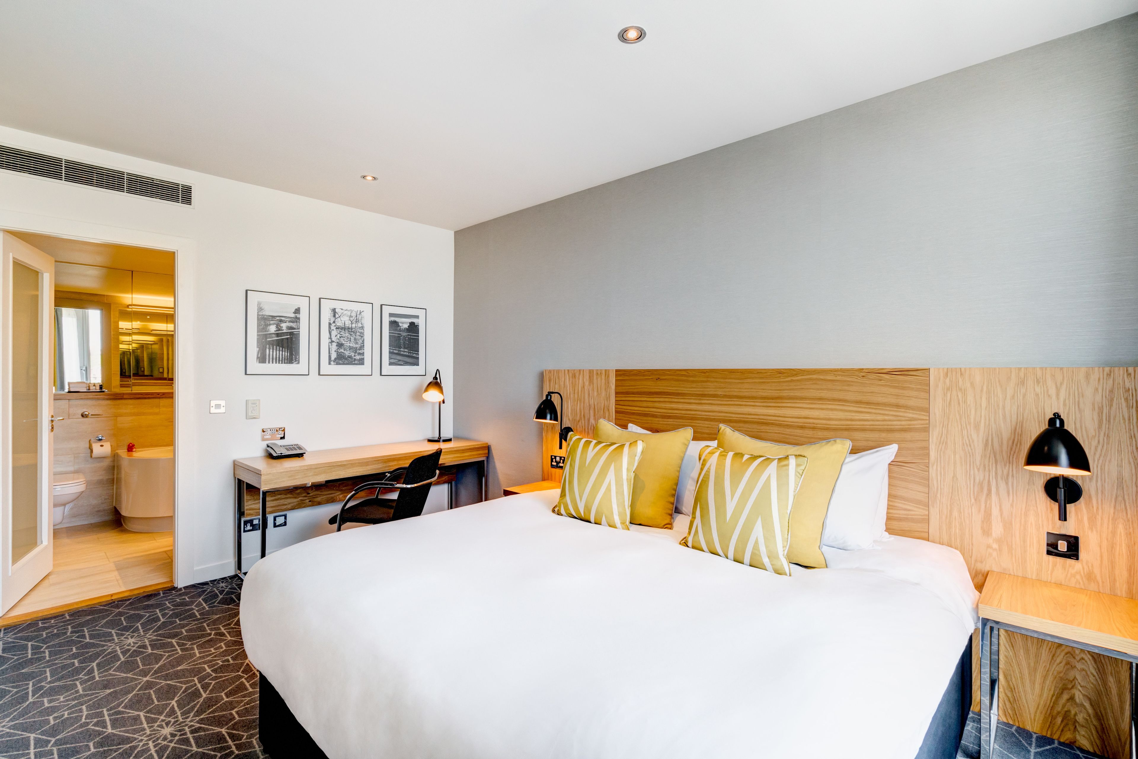 A hotel room with a large bed, a desk, and a chair. There are two wall-mounted lamps and a small table with a lamp.