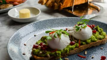 A plate with avocado toast topped with poached eggs and garnishes, a bowl of butter, and a basket of bread.