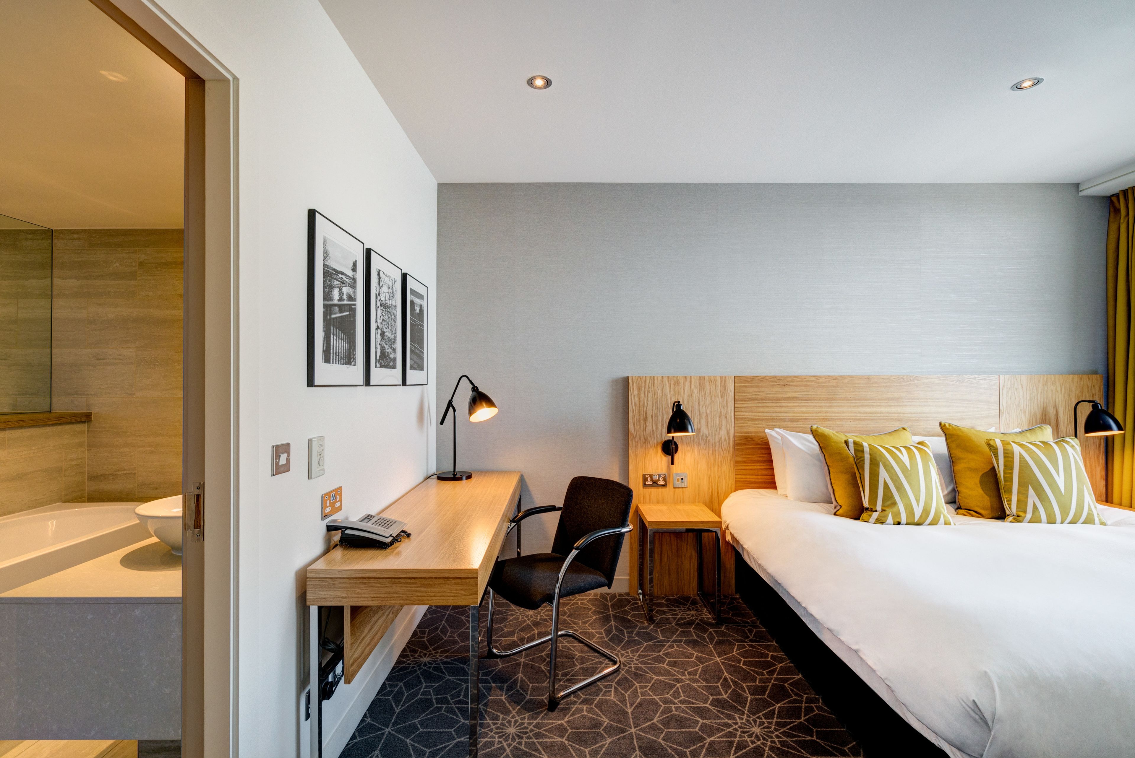 Une chambre d’hôtel moderne, équipée d’un lit, d’un bureau, d’une chaise et d’une salle de bain visible à travers une porte ouverte.