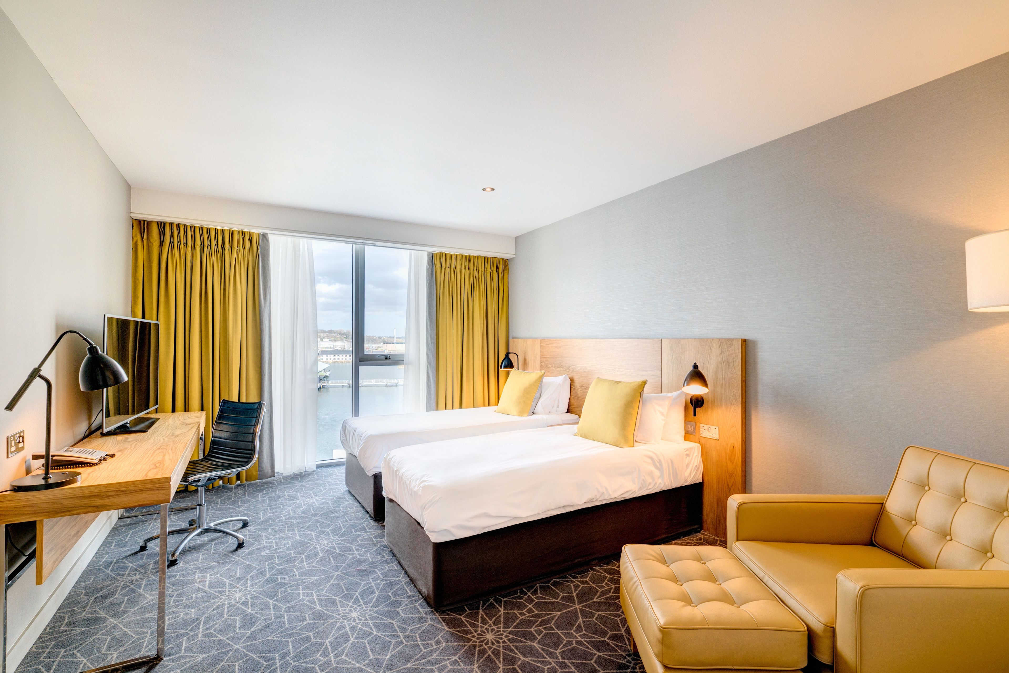 Une chambre d’hôtel avec deux lits, un bureau avec une chaise, un canapé et une grande fenêtre avec des rideaux.