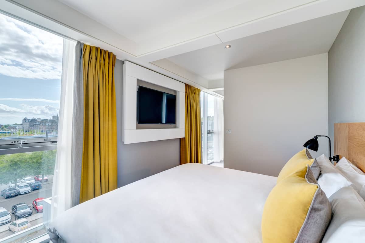 A modern hotel room with a large bed, a mounted TV, and a view of a cityscape through large windows.
