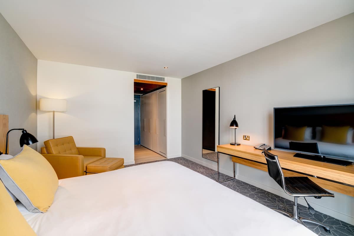A modern hotel room with a large bed, a desk with a flat-screen TV, a chair, and a lamp.