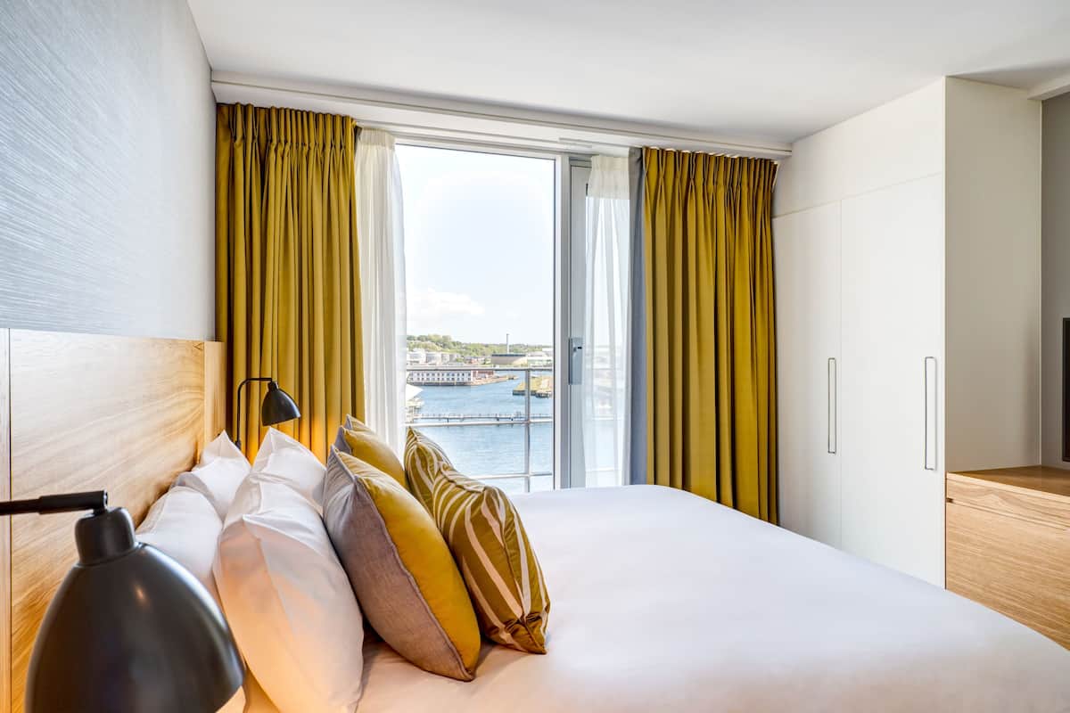 A hotel room with a large bed, a desk, and a view of a body of water through the windows.