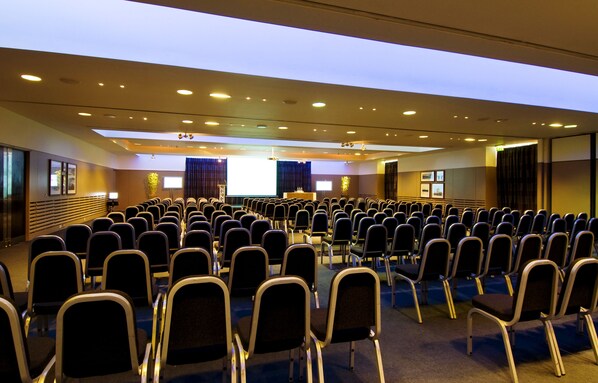 Un salón de conferencias con filas de sillas, un atril y una pantalla grande.