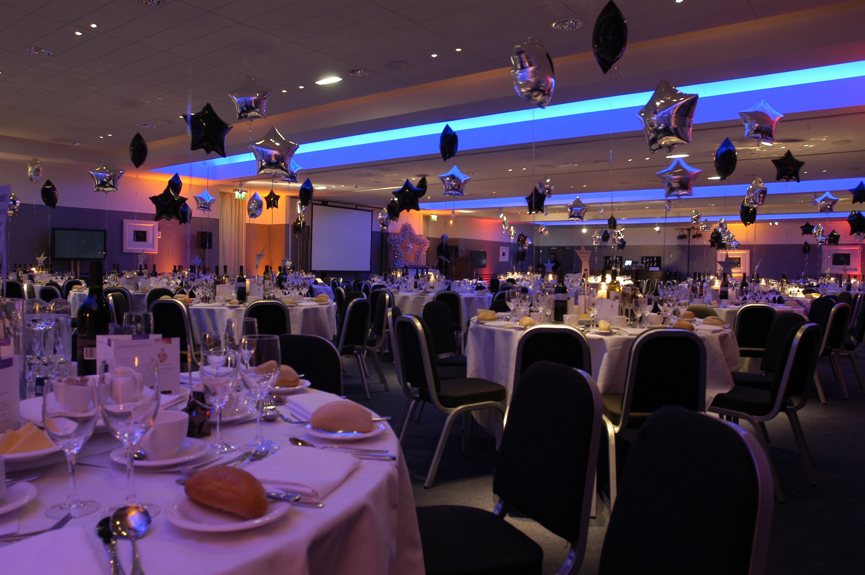 Une salle de banquet avec des tables rondes dressées pour un événement formel, comprenant la vaisselle et un écran de projection en arrière-plan.