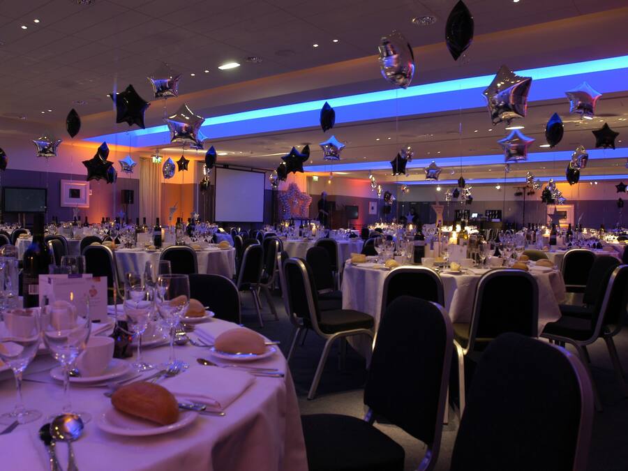 Une salle de banquet avec des tables rondes dressées pour un événement formel, comprenant la vaisselle et un écran de projection en arrière-plan.