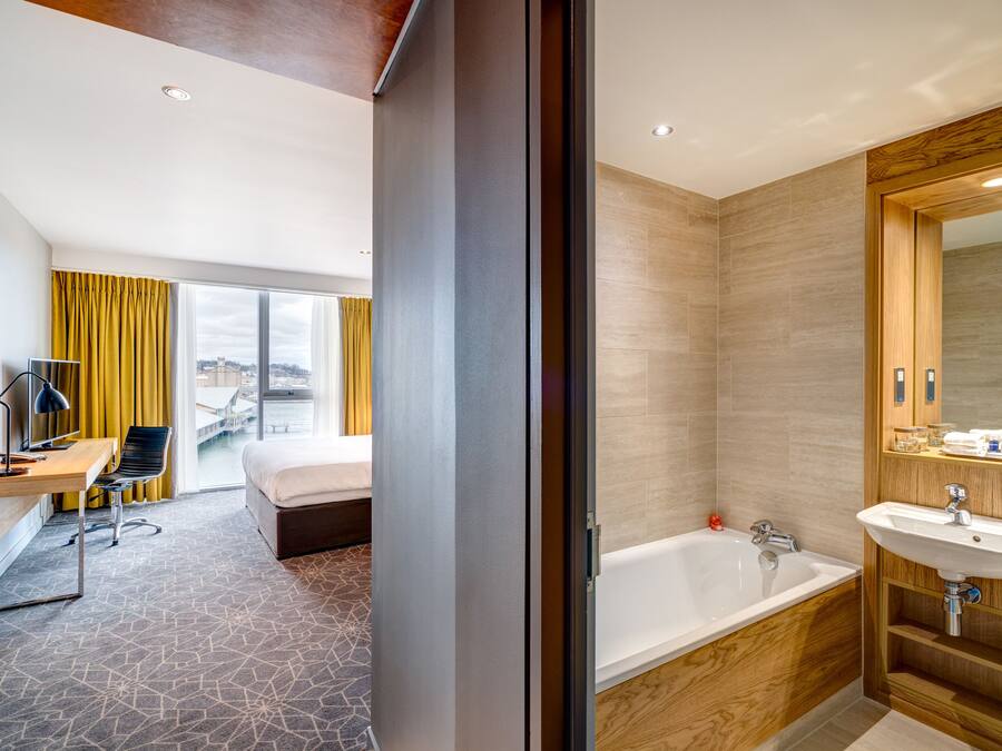 Ein modernes Hotelzimmer mit einem großen Bett, einem Schreibtisch mit Stuhl, einem Laptop und einem Fenster mit Vorhängen.