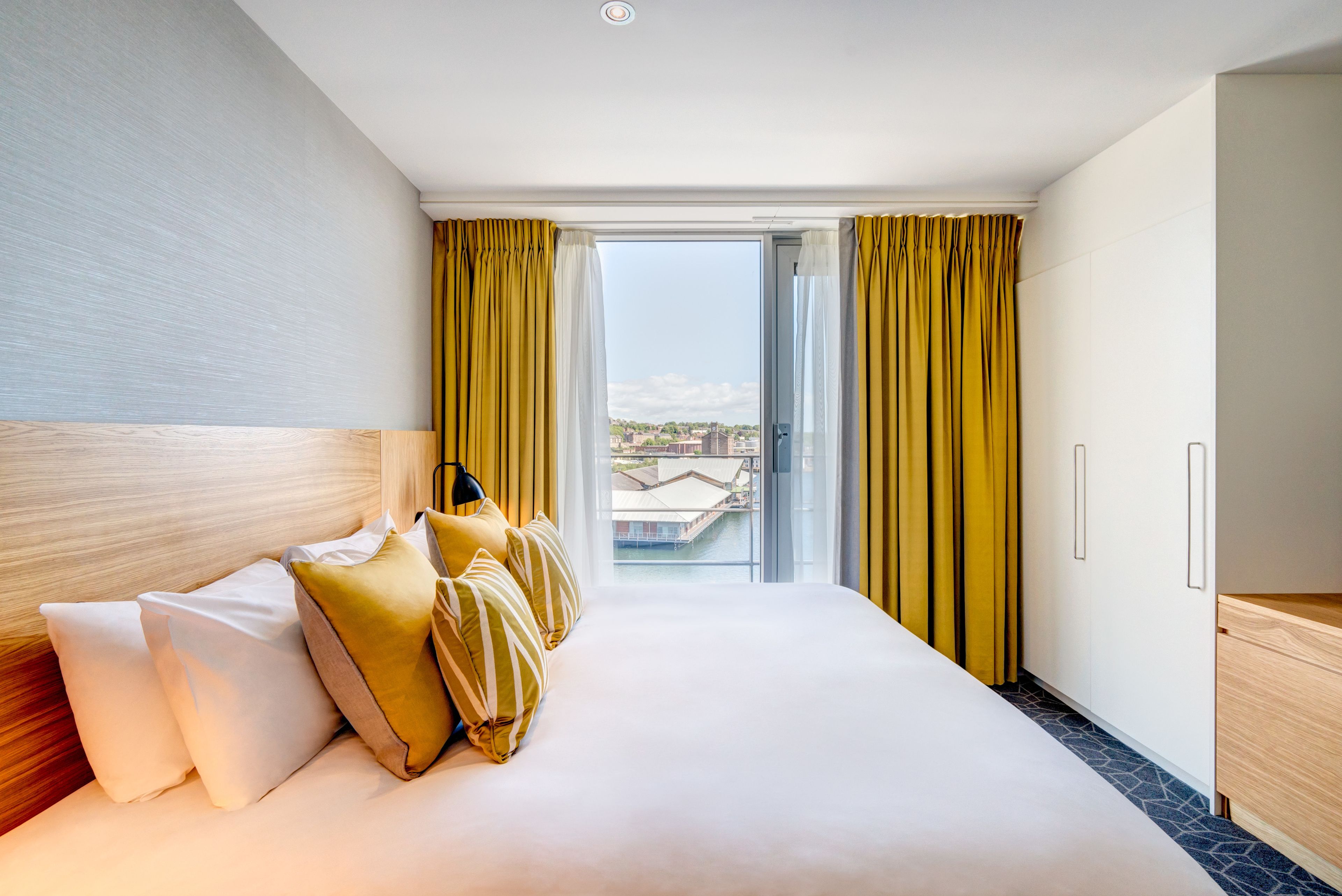 A hotel room with a large bed, a wooden headboard, a view through a sliding door, and a balcony with yellow curtains.