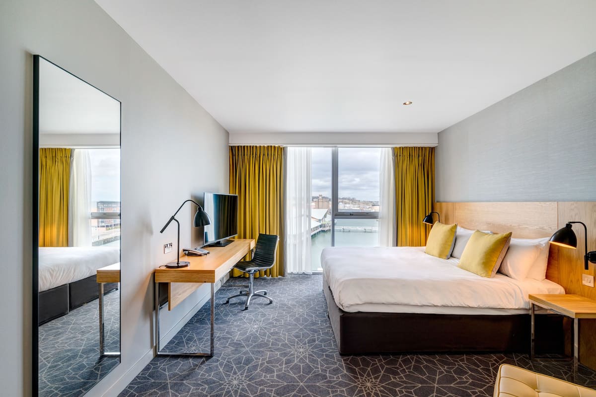 A modern hotel room with a large bed, a desk with a computer, a chair, and a large mirror.