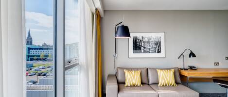 A modern hotel room with a large window, a sofa with patterned cushions, a desk, and a framed artwork on the wall.