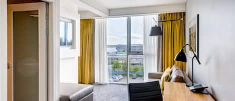 Una habitación de hotel moderna con un amplio ventanal corredero que da a un balcón, un sofá, una mesita de madera con lámpara y vistas a la ciudad.