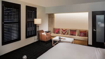Hotelkamer met een bed, een bank, een stoel, een lamp en twee ramen met jaloezieën.