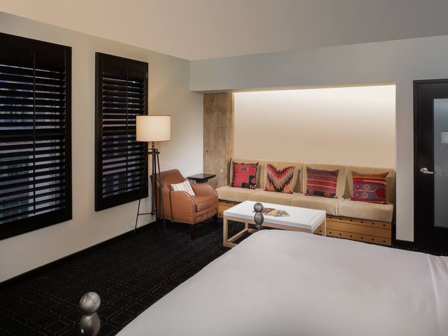 Hotelkamer met een bed, een bank, een stoel, een lamp en twee ramen met jaloezieën.