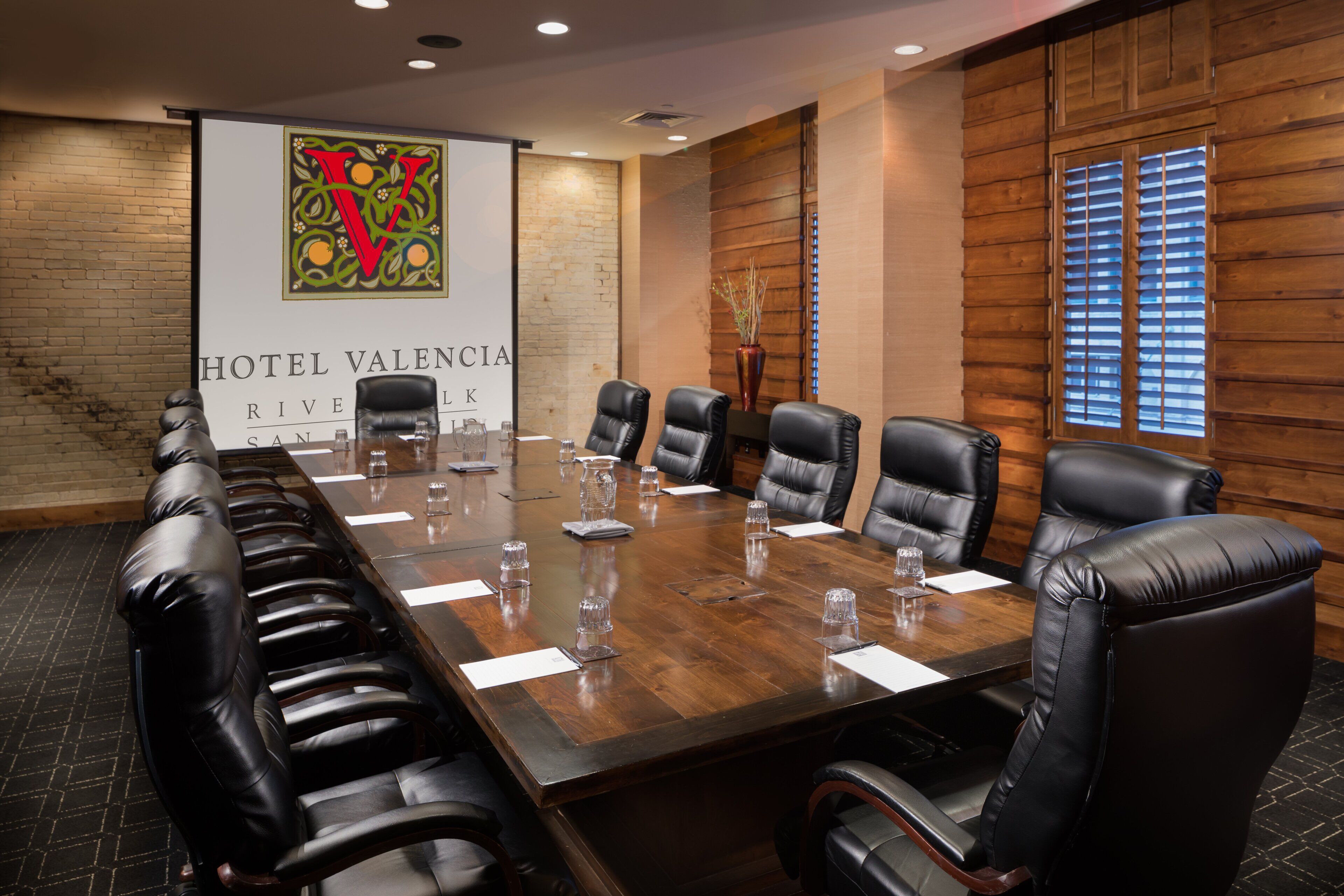 A conference room with a long table, black leather chairs, and a banner reading "Hotel Valencia River San Luis".