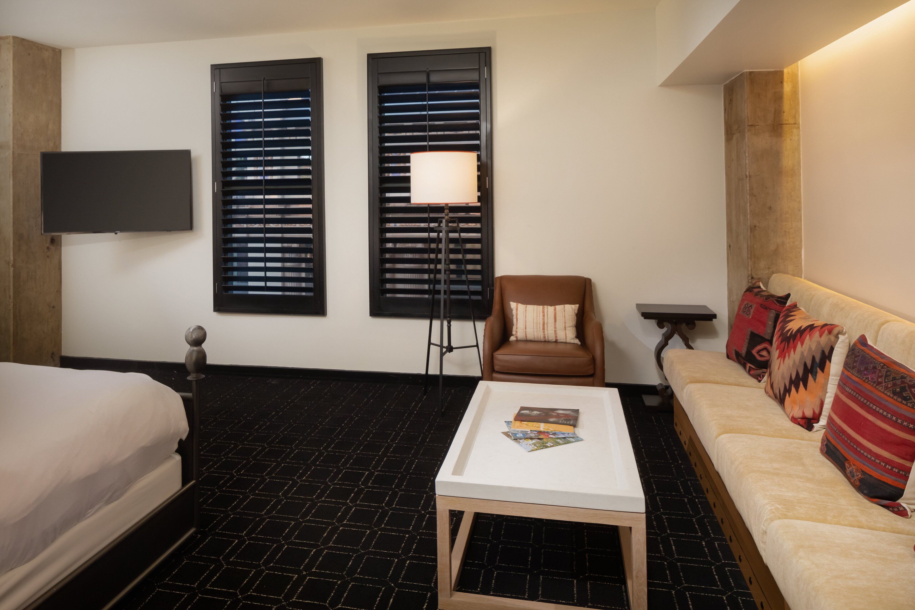 A hotel room with a bed, a sofa, a coffee table, a chair, a TV, and two windows with blinds.